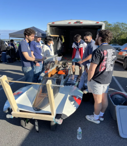 Group of students working on a small race boat next to an open trailer in a parking lot.